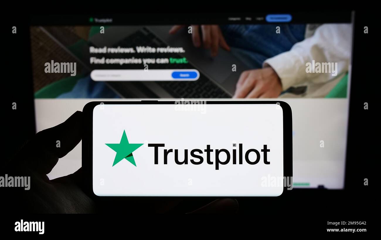 Persona che tiene uno smartphone con il logo della società di revisione della piattaforma Trustpilot Group plc sullo schermo di fronte al sito Web. Messa a fuoco sul display del telefono. Foto Stock