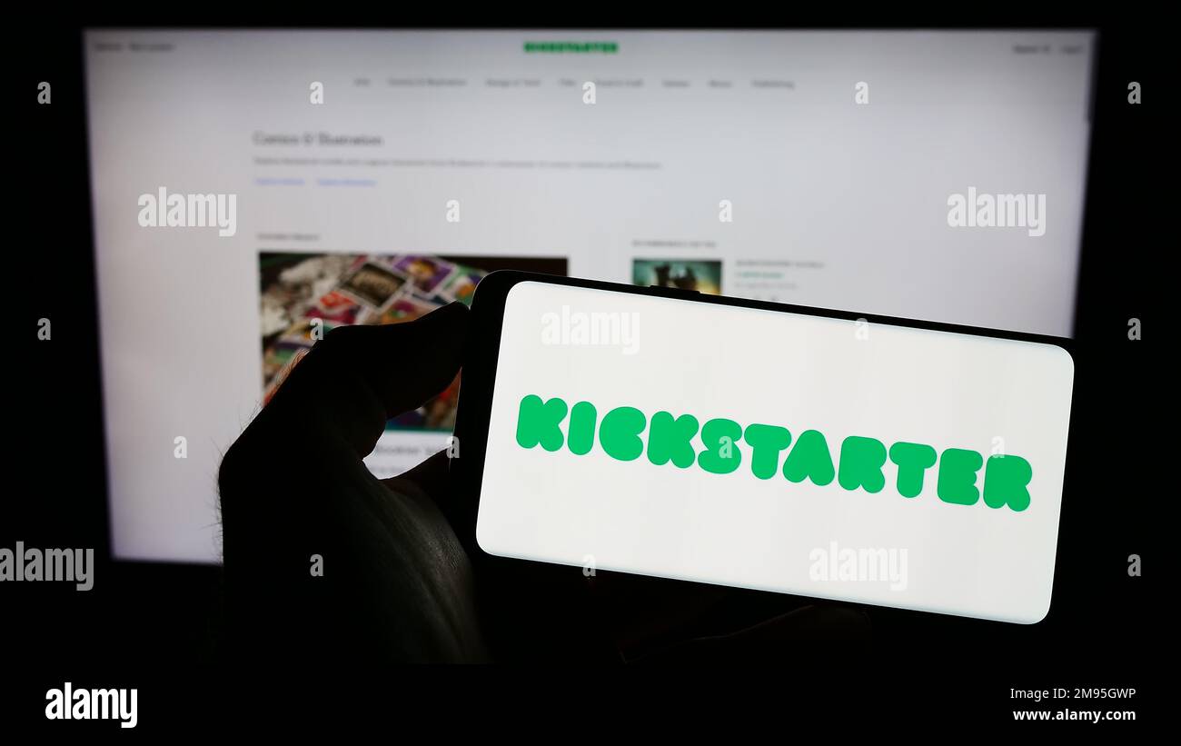 Persona che tiene il cellulare con il logo della piattaforma di crowdfunding US Kickstarter PBC sullo schermo di fronte alla pagina web aziendale. Messa a fuoco sul display del telefono. Foto Stock