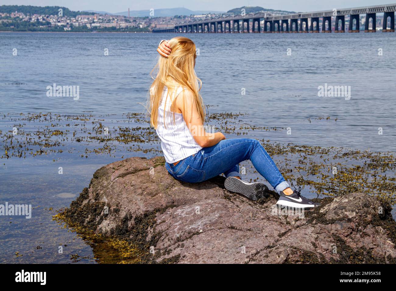 Rhianna Martin, una bella donna bionda, si siede sulle rocce ammirando la vista sulla spiaggia di Wormit nella contea di Fife, Scozia, Regno Unito Foto Stock