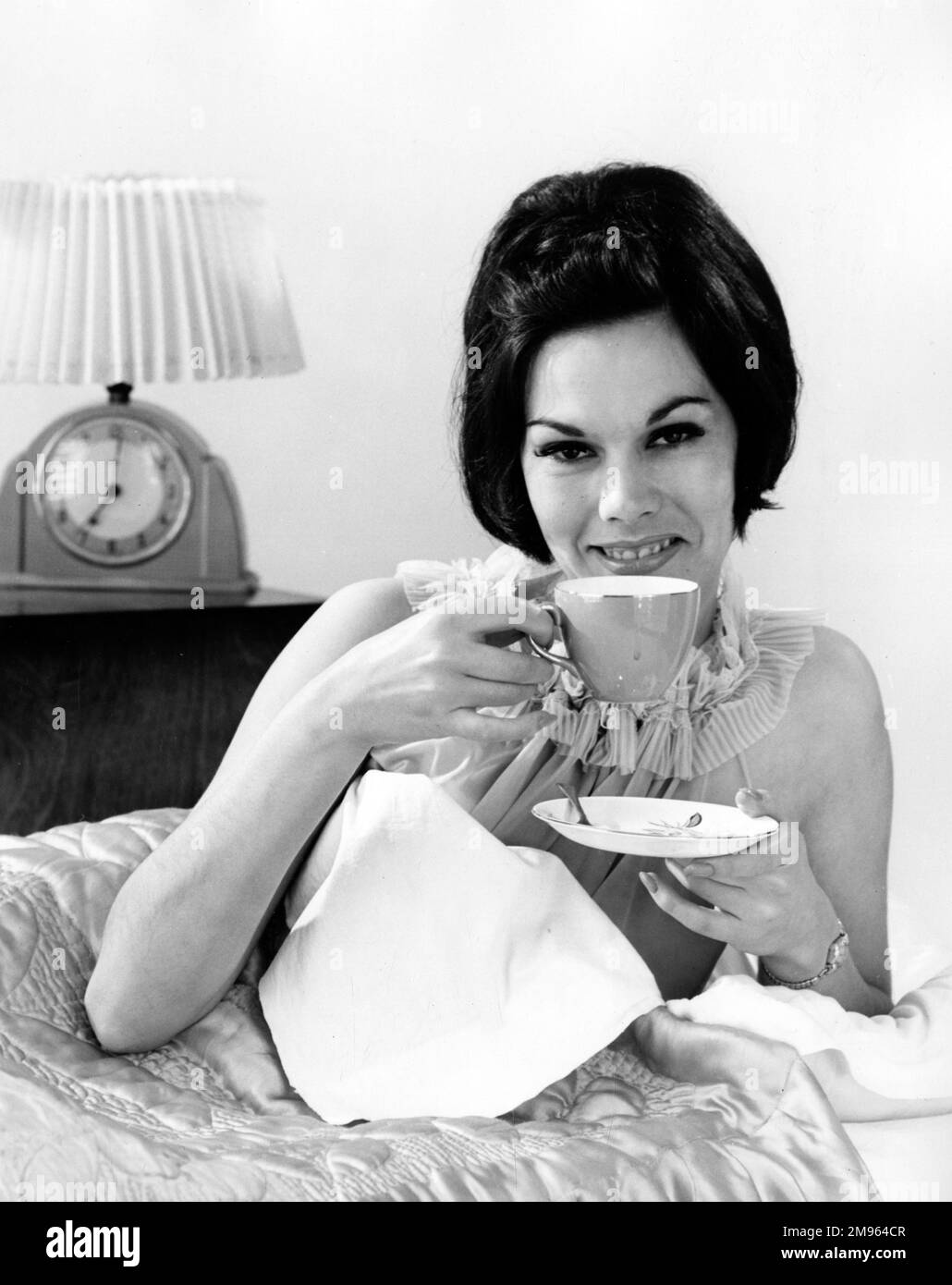 Una donna si trova a letto con una tazza di tè. È 7:01 così abbondanza di tempo per una bugia dentro. Foto Stock