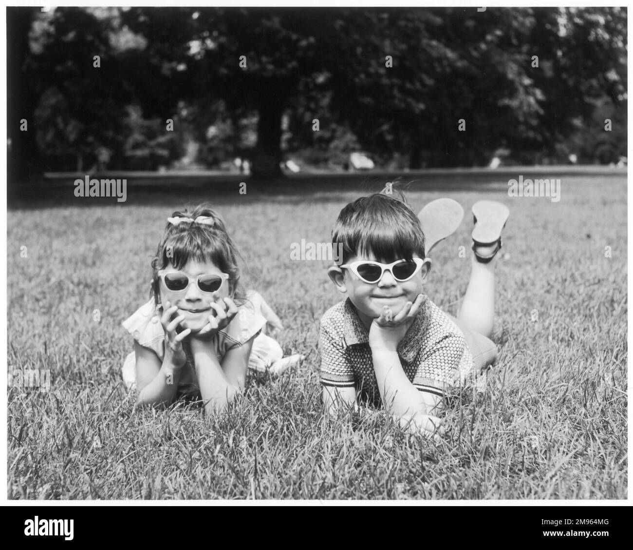 Due bambini giacciono sull'erba in un parco, ognuno con un paio di occhiali da sole. Foto Stock