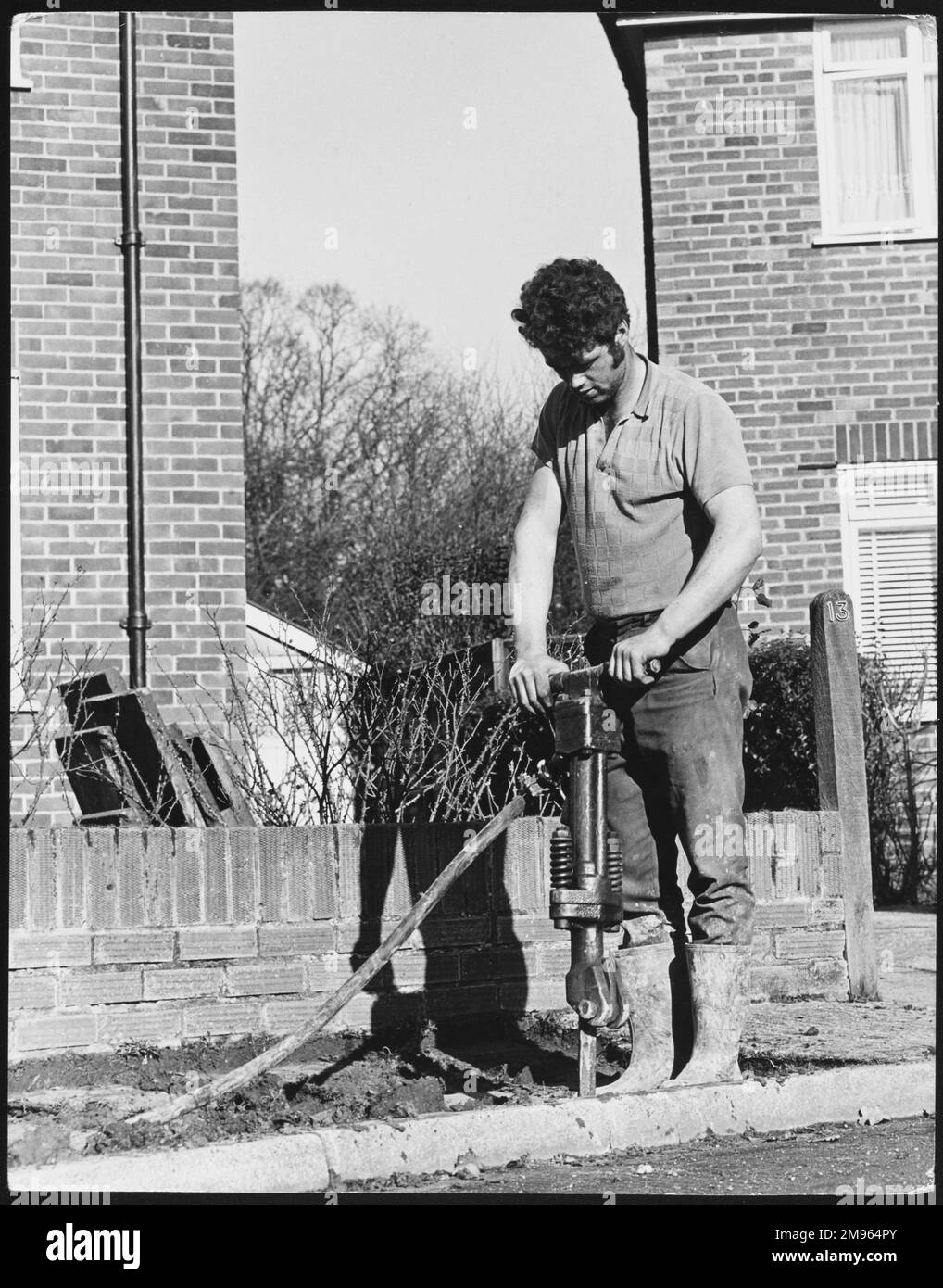 Un uomo con capelli ricci e scuri che usa un trapano pneumatico per scavare una vecchia pavimentazione. Foto Stock