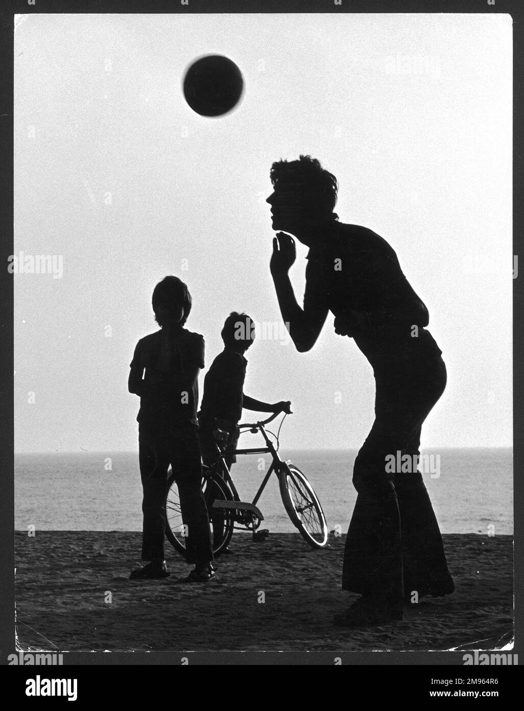 Silhouette di un adolescente che dirige un calcio su una spiaggia, guardato da due ragazzi più piccoli, uno che tiene la bicicletta. Foto Stock