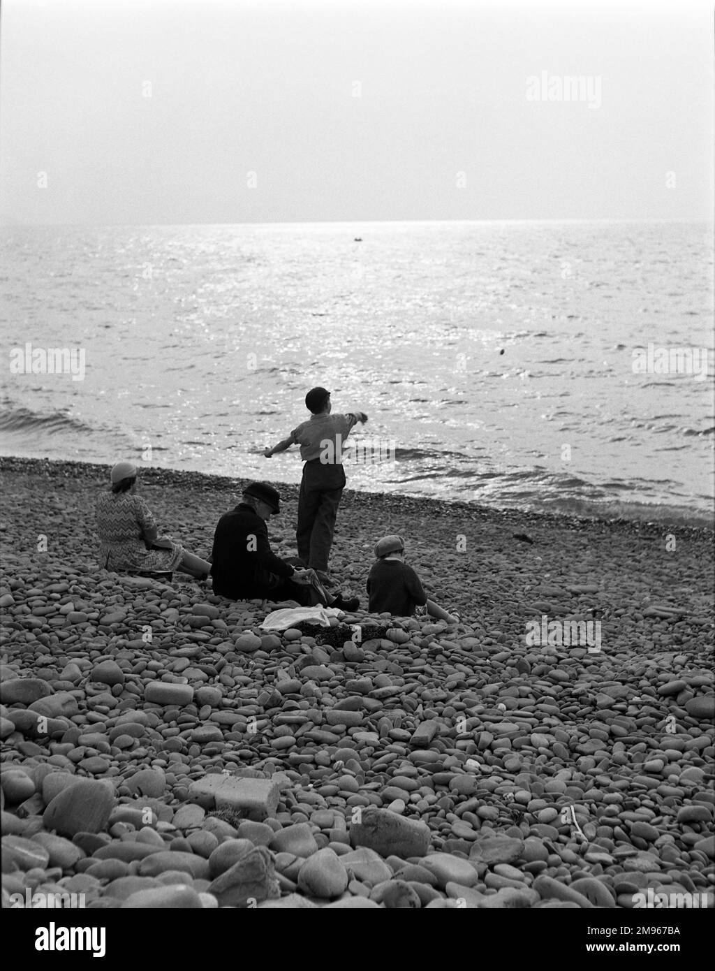Skimming pietre. Un ragazzino, in piedi accanto alla sua famiglia seduto, sbatte pietre piatte nel mare dalla spiaggia di ciottoli, cercando di farli scorrere attraverso la superficie alla luce della sera. Fotografia di Norman Synge Waller Budd Foto Stock