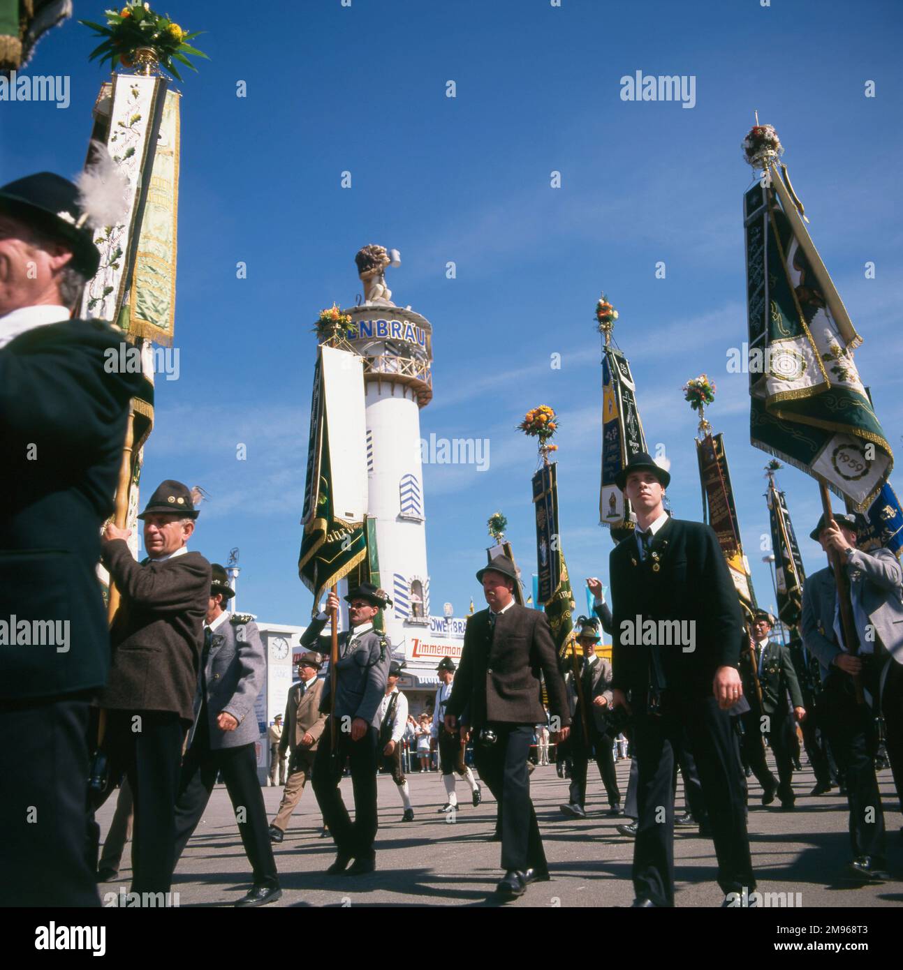 Scena all'Oktoberfest di Monaco, Germania, con persone in costume popolare che marciano con bandiere, e la torre della Lowenbrau Brewery sullo sfondo. Foto Stock