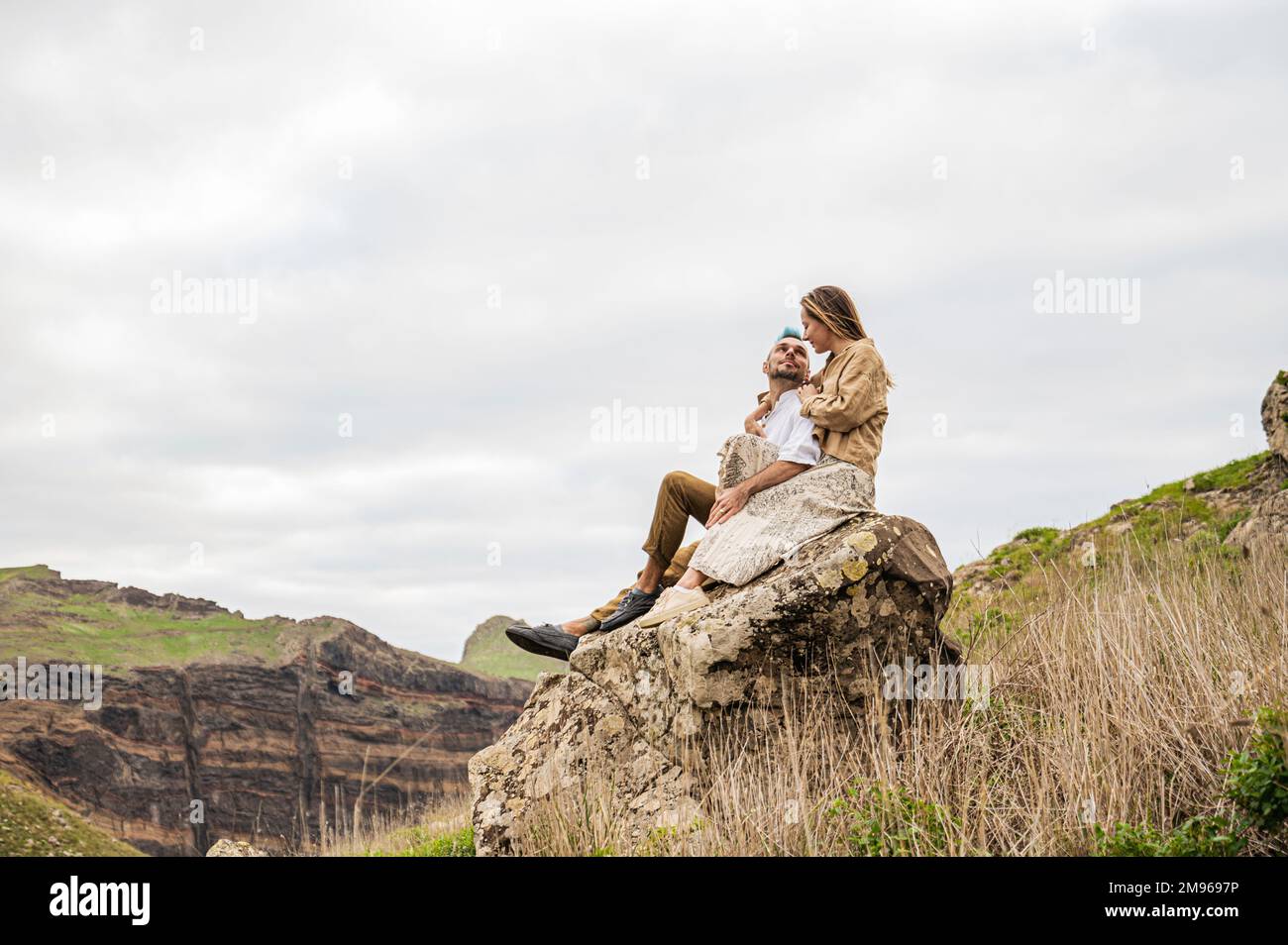 Una giovane coppia siede su una roccia circondata da colline e rocce sullo sfondo Foto Stock
