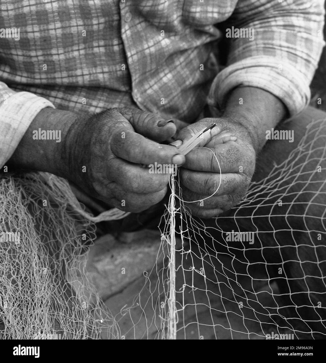 Un primo piano delle mani di un pescatore che indossava fatica, mentre si siede a mendersi una rete. Foto Stock
