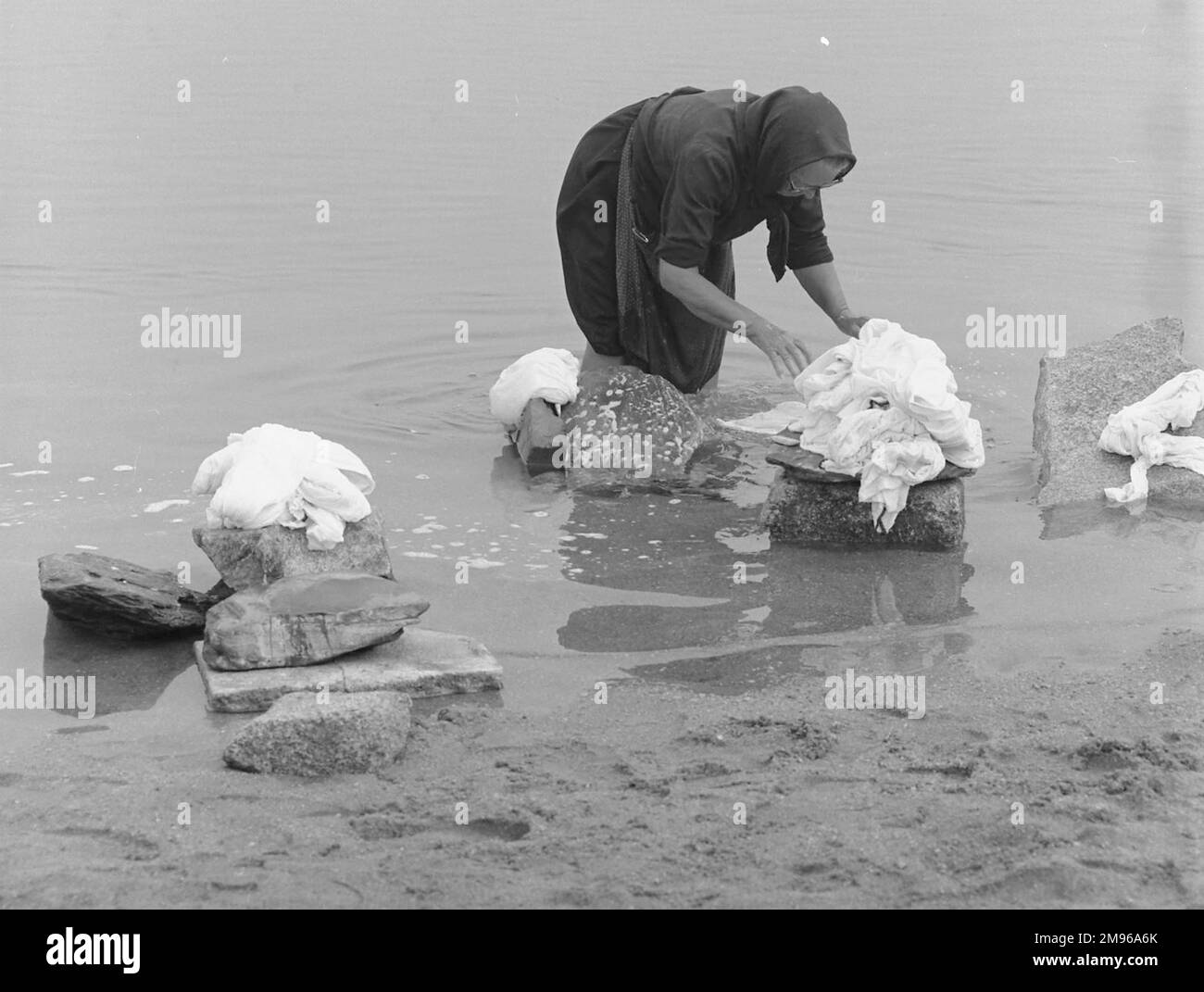 Una donna che sta in piedi ginocchio in un fiume, ordinando gli oggetti per il lavaggio che ha messo su varie rocce. Foto Stock Una donna che sta in piedi ginocchio in un fiume, ordinando gli oggetti per il lavaggio che ha messo su varie rocce. Foto Stock