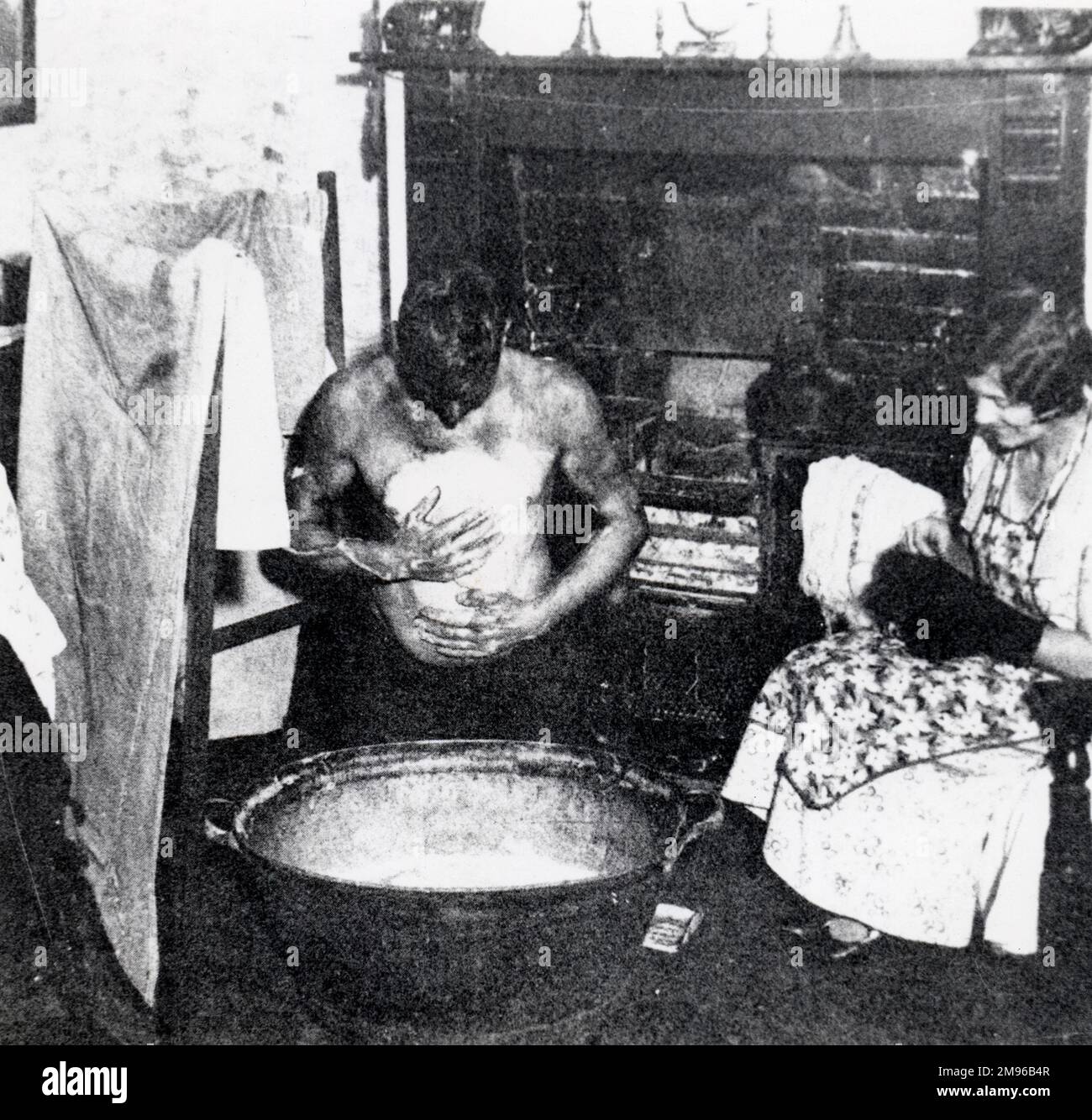 Un minatore del Galles del Sud si lava in un bagno di latta di fronte al fuoco, mentre sua moglie siede calze mendenti. Foto Stock