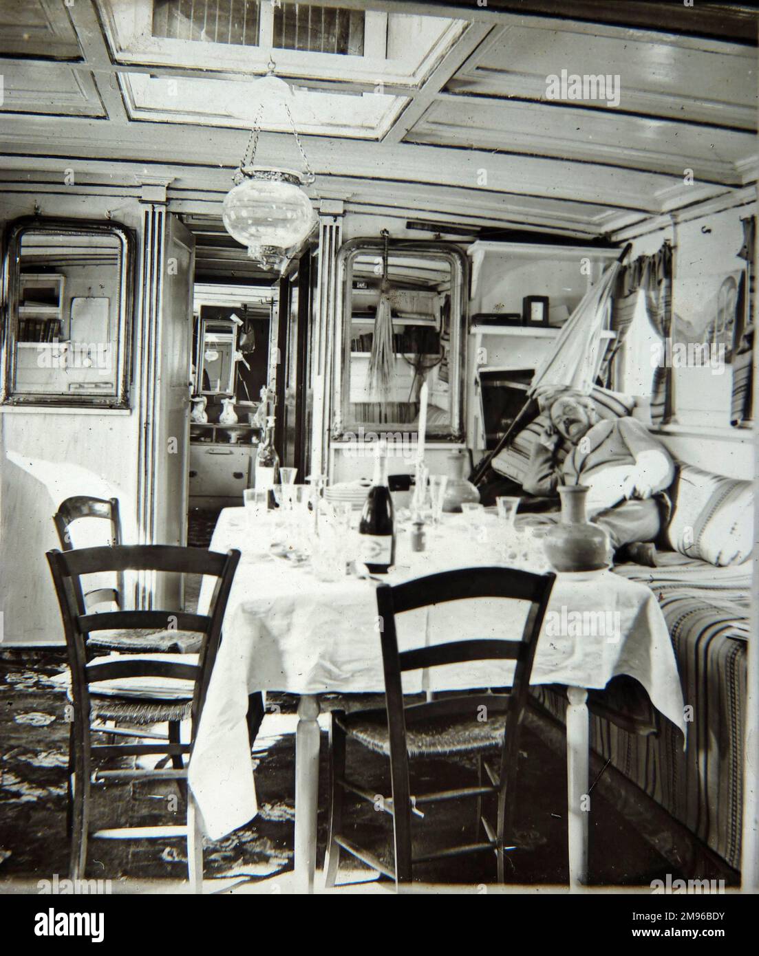 Vista dell'interno di una barca edoardiana. Un tavolo è impostato, ma forse più per bere che mangiare, con una bottiglia e diversi bicchieri. Ci sono tre sedie verticali con sedili in vimini. Un uomo giace sui cuscini di una panca (a destra), guardando direttamente la telecamera. Una stretta porta aperta mostra che c'è un'altra piccola stanza dietro. Foto Stock