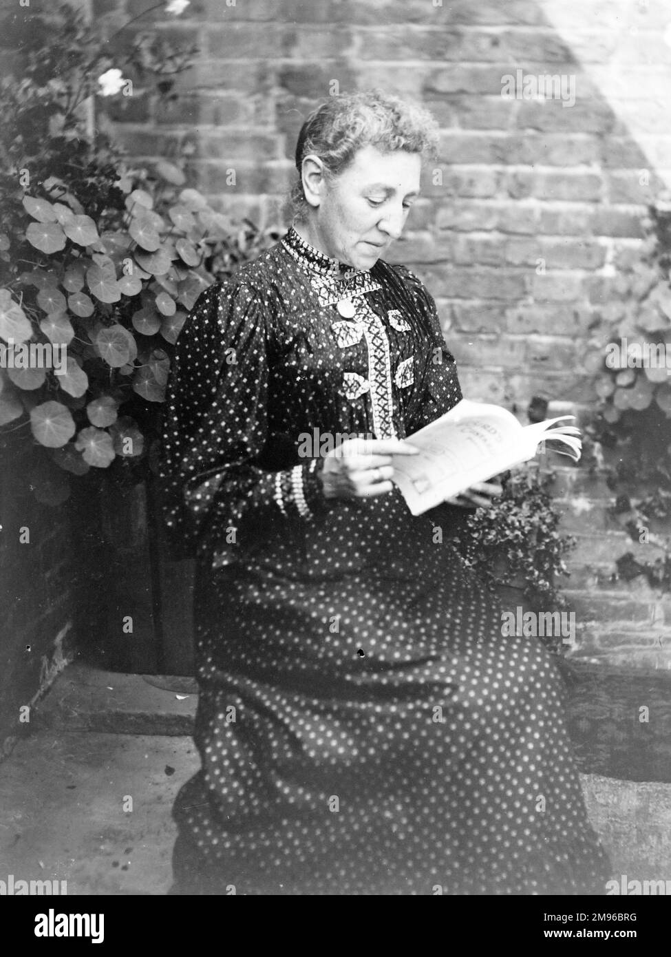 Una donna di mezza età seduta a leggere in un giardino, Galles centrale. Indossa un abito a tutta lunghezza macchiato. Foto Stock