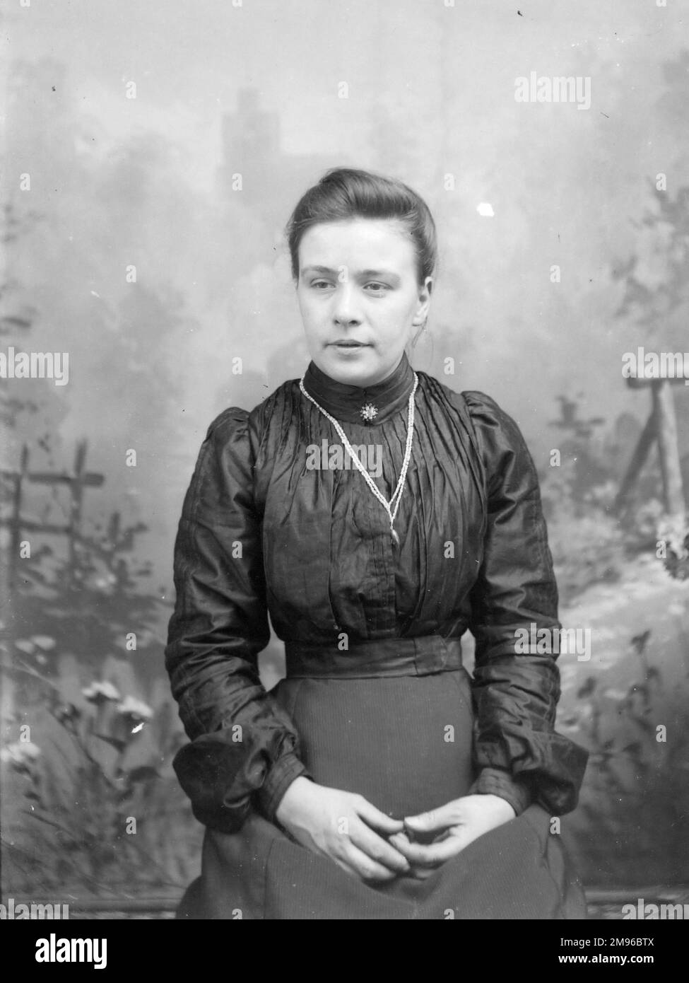 Una giovane donna edoardiana in abiti scuri posa per la sua foto nello studio del fotografo, Mid Wales. È seduta, con una scena di paese dipinta sullo sfondo dietro di lei. Foto Stock