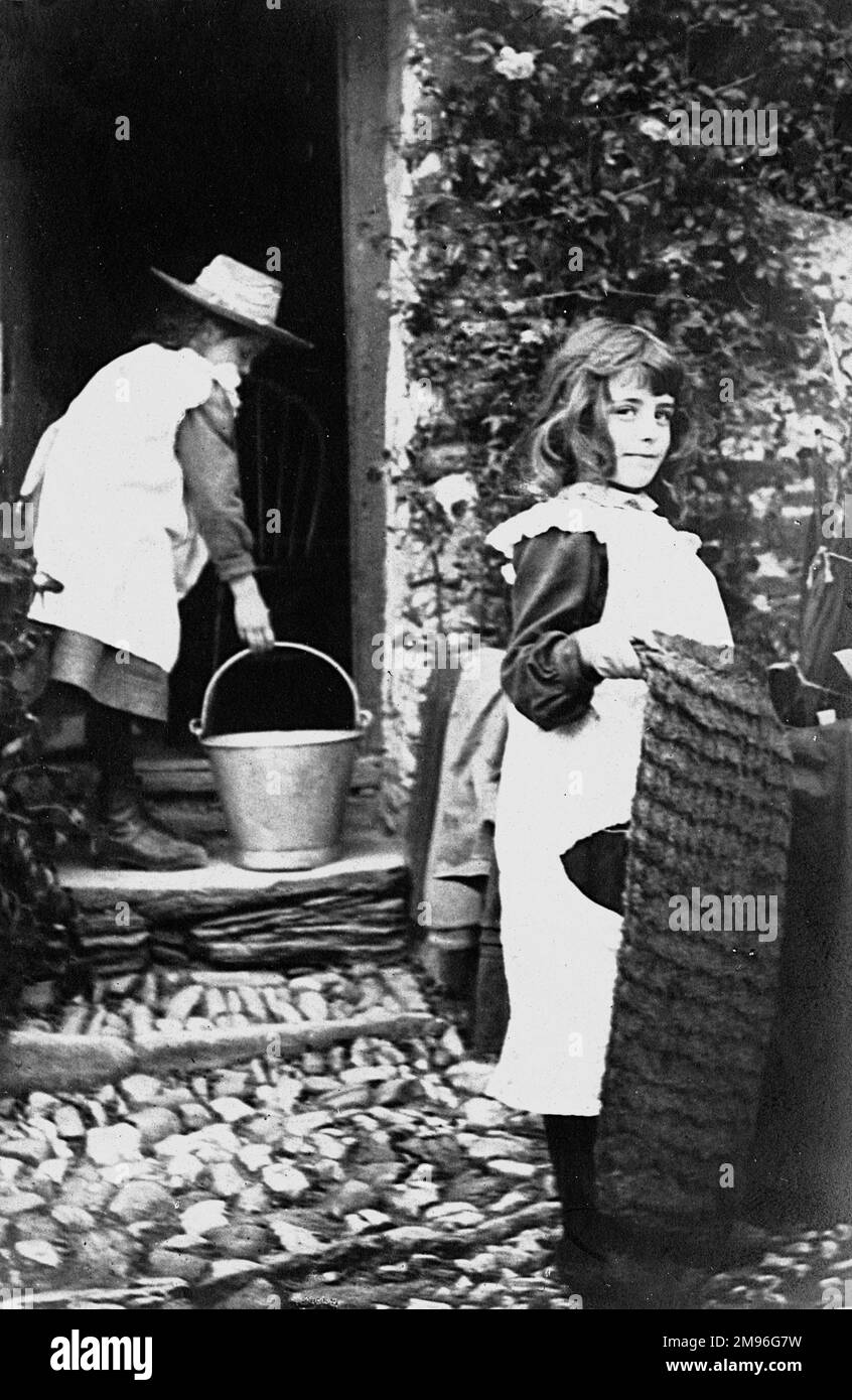 Due ragazze aiutano la loro madre con il lavoro di casa; una tiene un secchio di acqua, l'altra scuote giù una stuoia. Foto Stock