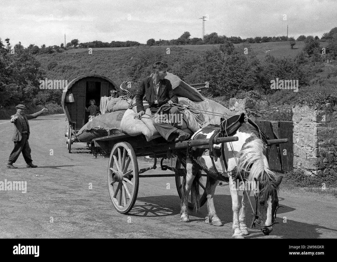 Una strada di campagna con un giovane alla guida di un cavallo e un carro. Una carovana zingara segue dietro. Foto Stock