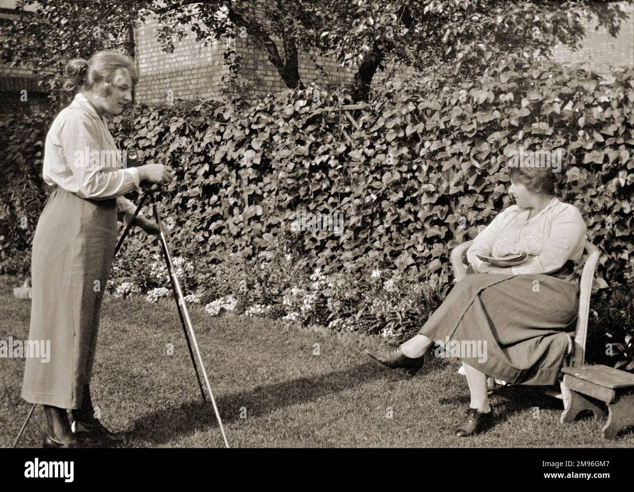 Due donne in giardino. Una donna di mezza età è seduta, pronta per essere fotografata da una donna più giovane che ha una macchina fotografica su un treppiede. Foto Stock