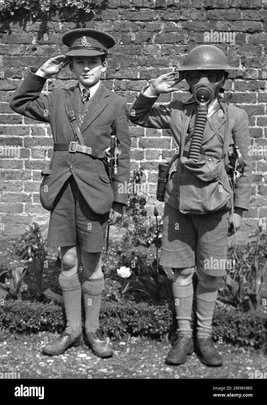 Due ragazzi salutano, probabilmente durante la seconda guerra mondiale. Quello a destra indossa un casco di latta e una maschera a gas. Quello a sinistra indossa un berretto di artiglieria reale e una spada cerimoniale. Foto Stock