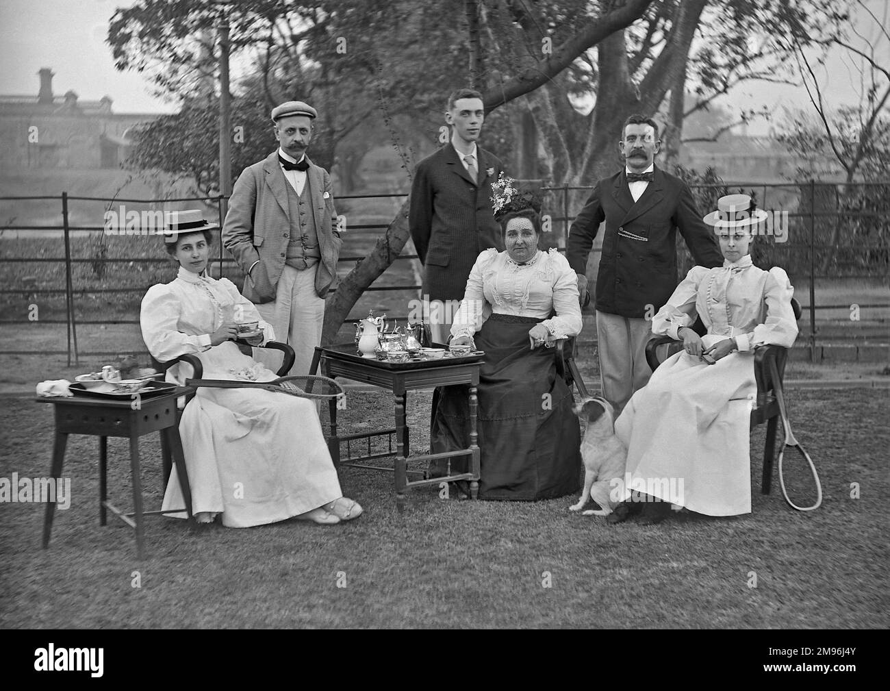 Tre uomini e tre donne prendono il tè in un giardino. Un cane piccolo guarda in su espettantly. Le due giovani donne hanno racchette da tennis. Foto Stock