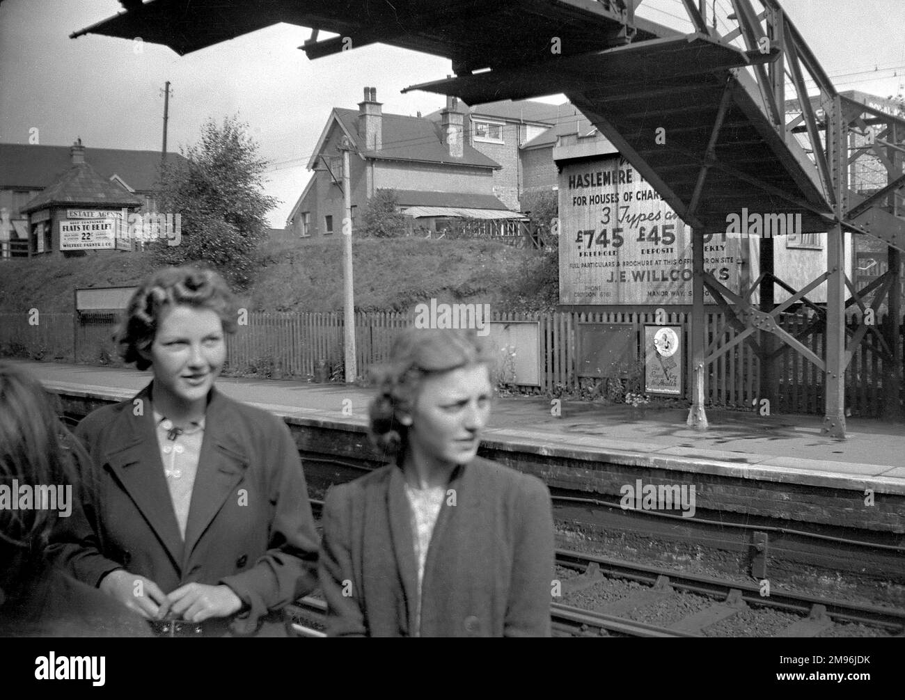 Due giovani donne in una stazione ferroviaria nel sud di Londra. Un annuncio sulla piattaforma opposta è per proprietà, case di carattere per £745 freehold. Un altro segno fa pubblicità agli appartamenti auto-contenuti per lasciare. Foto Stock
