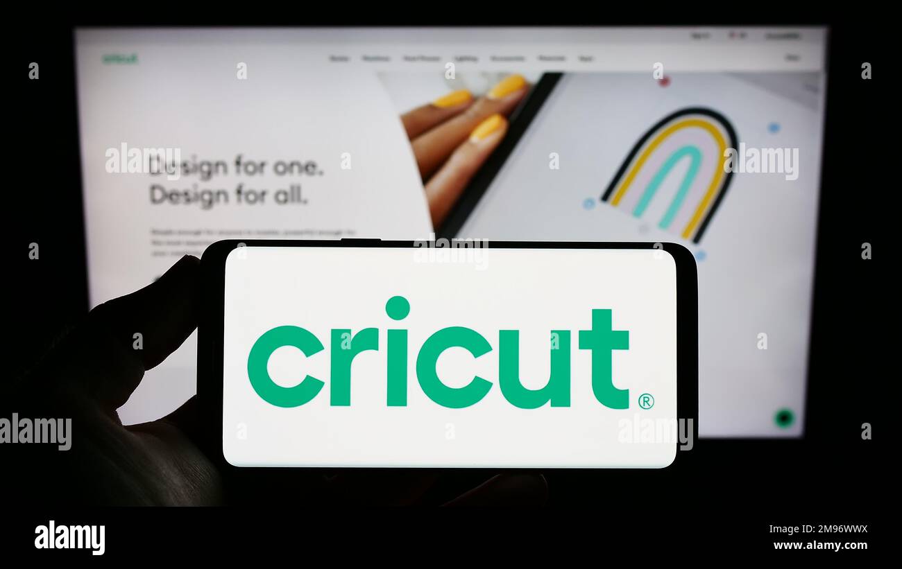 Persona che tiene in mano uno smartphone con il logo dell'azienda statunitense Cricut Inc. Che produce plotter da taglio sullo schermo di fronte al sito Web. Messa a fuoco sul display del telefono. Foto Stock
