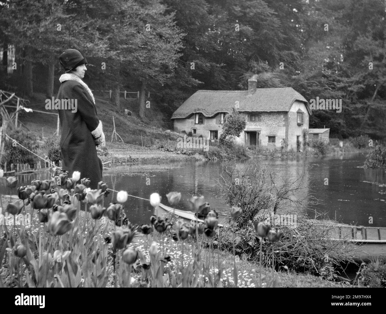 Donna in piedi accanto a un lago con tulipani in primo piano e un cottage di paglia in lontananza. Foto Stock