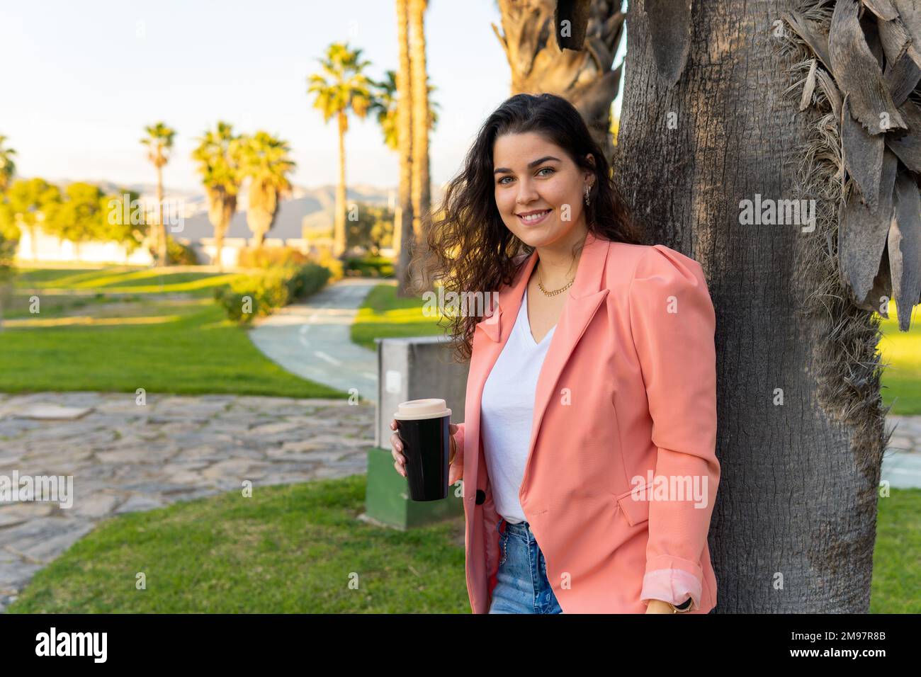 Giovane ragazza latina ben vestita che tiene il suo bicchiere di caffè appoggiato contro un albero in un parco all'aperto. Foto Stock