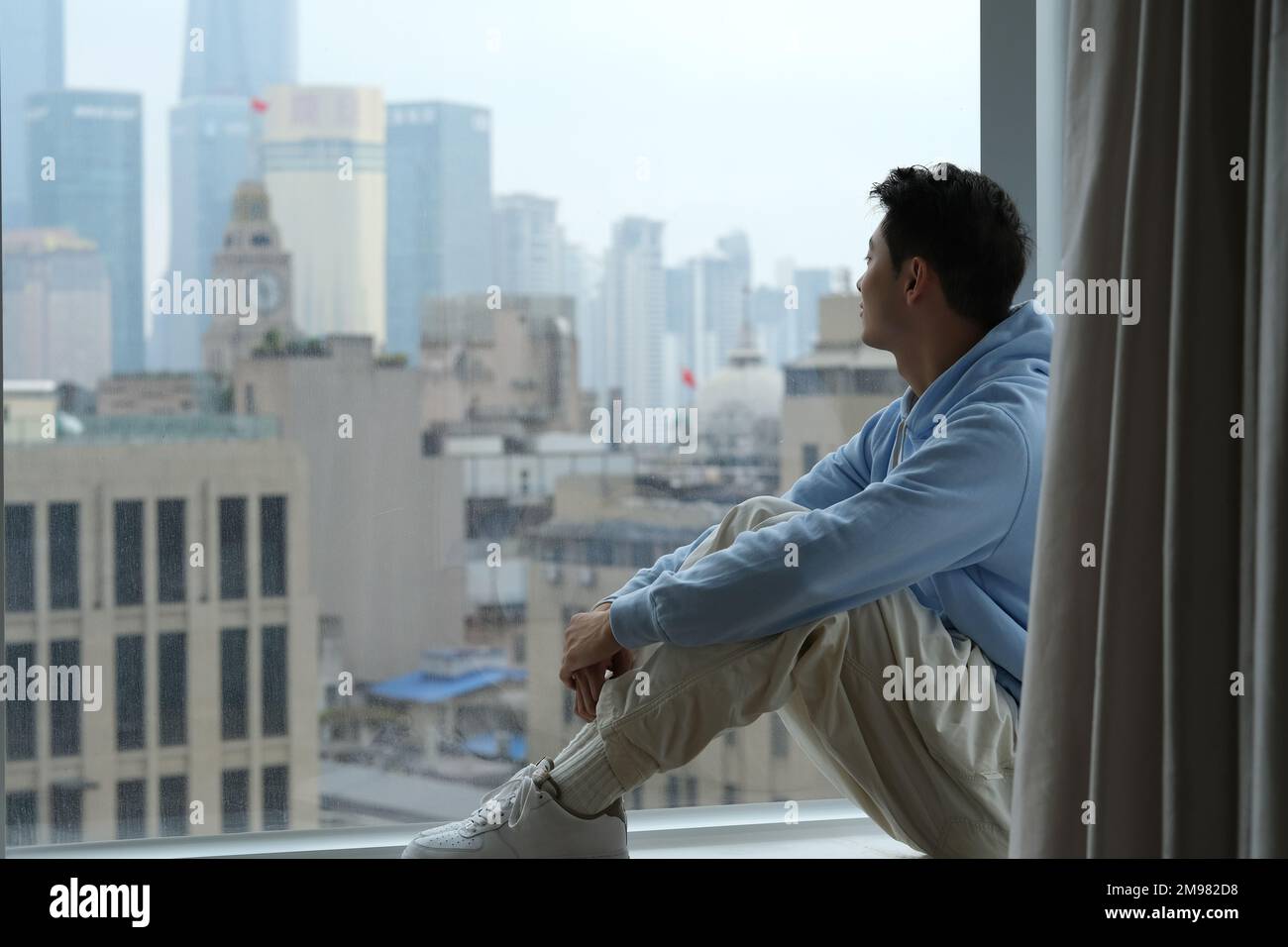 Un giovane cinese solitario siede al davanzale della finestra, guarda fuori dalla finestra per vedere gli edifici urbani di Shanghai, Cina Foto Stock