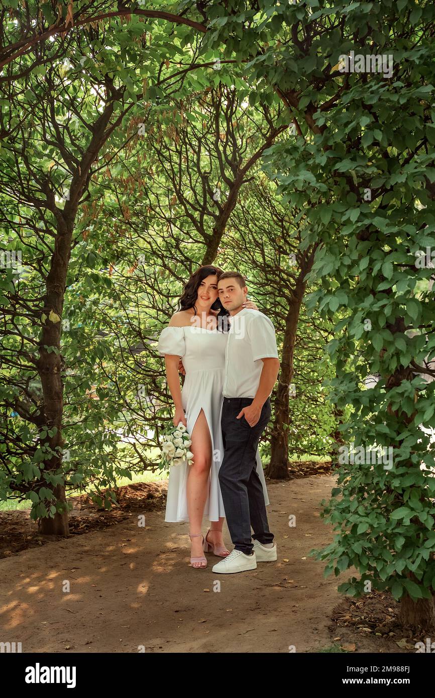 Ritratto di sposa felice e sposo sotto l'arco verde fiorito primo piano. Una coppia gioiosa nel giardino primaverile, una ragazza con un bouquet di bianco lussureggiante Foto Stock