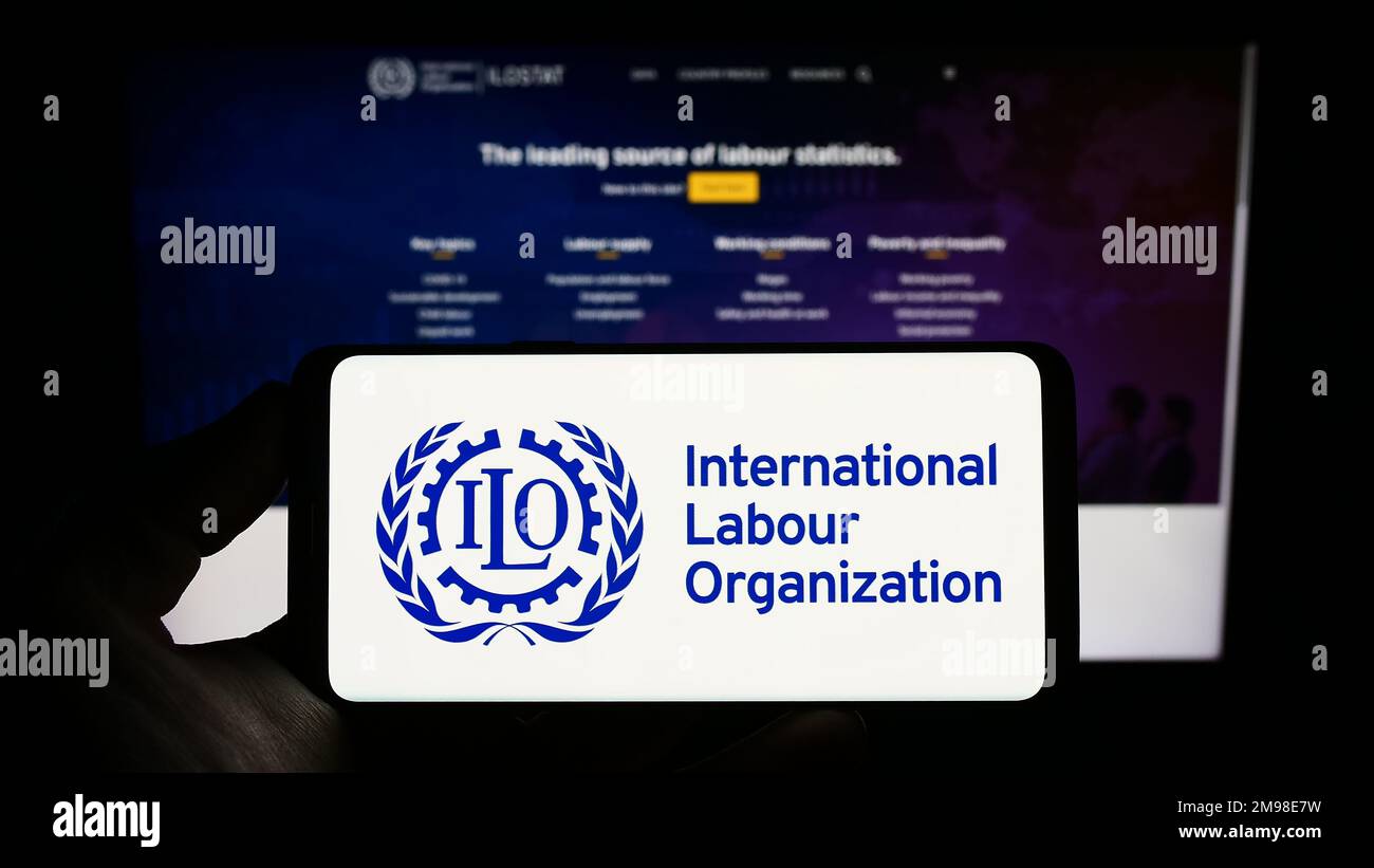 Persona che tiene in mano uno smartphone con il logo dell'Organizzazione Internazionale del lavoro (ILO) dell'agenzia delle Nazioni Unite sullo schermo di fronte al sito Web. Messa a fuoco sul display del telefono. Foto Stock