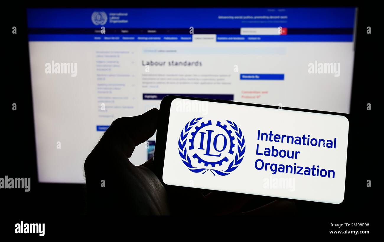 Persona che tiene il cellulare con il logo dell'agenzia ONU International Labour Organization (ILO) sullo schermo di fronte alla pagina web. Messa a fuoco sul display del telefono. Foto Stock