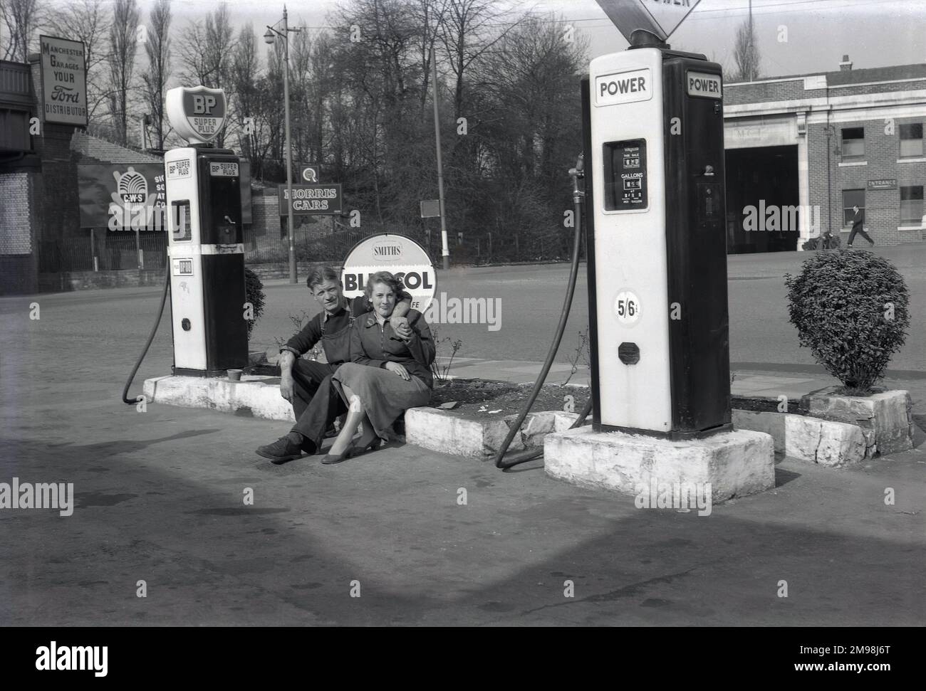 1950s, storico, meccanico maschile e impiegato femminile dell'Harbood Motor Co seduto per una foto da pompe di benzina per Power e BP carburante sul piazzale del garage, Wilmslow Rd, Parrs Wood, Didsbury, Manchester, Inghilterra, Regno Unito. Il prezzo della benzina Power è di 5/6d (5s 6d) un gallone, costoso, come era il momento della crisi di Suez. Un grande deposito di autobus che serve la città di Manchester è dall'altra parte della strada. Un cartello per Morris Cars può essere visto all'ingresso del ponte ferroviario, con la stazione ferroviaria East Didsbury & Parrs Wood nelle vicinanze. Foto Stock