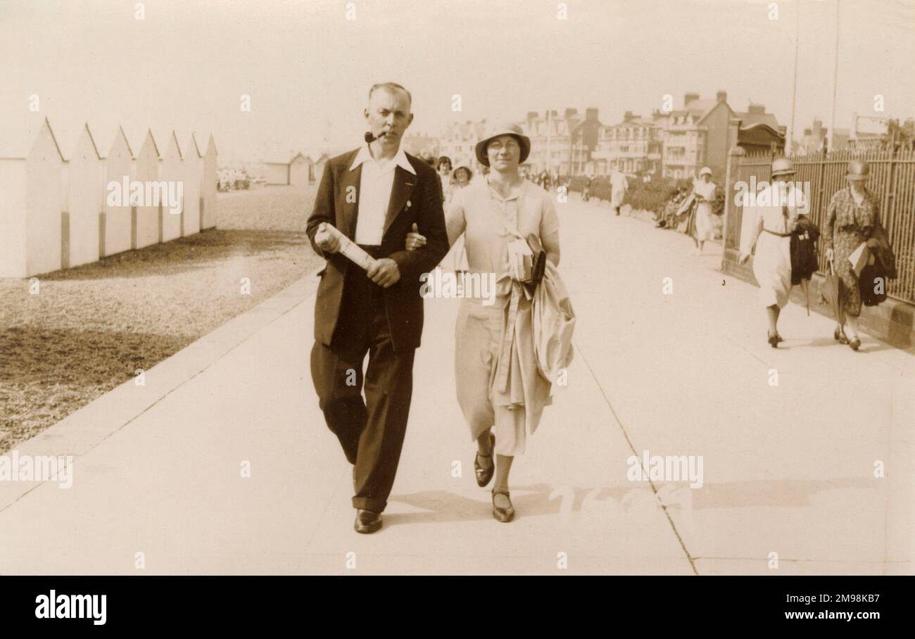 Coppia in vacanza passeggiata lungo la passeggiata a Felixstowe, Suffolk. I badge sulla sua giacca suggeriscono che forse è un railwayman e sua moglie sta portando un paio di romanzi a mentre via le ore residenti nella sua sedia a sdraio... Foto Stock
