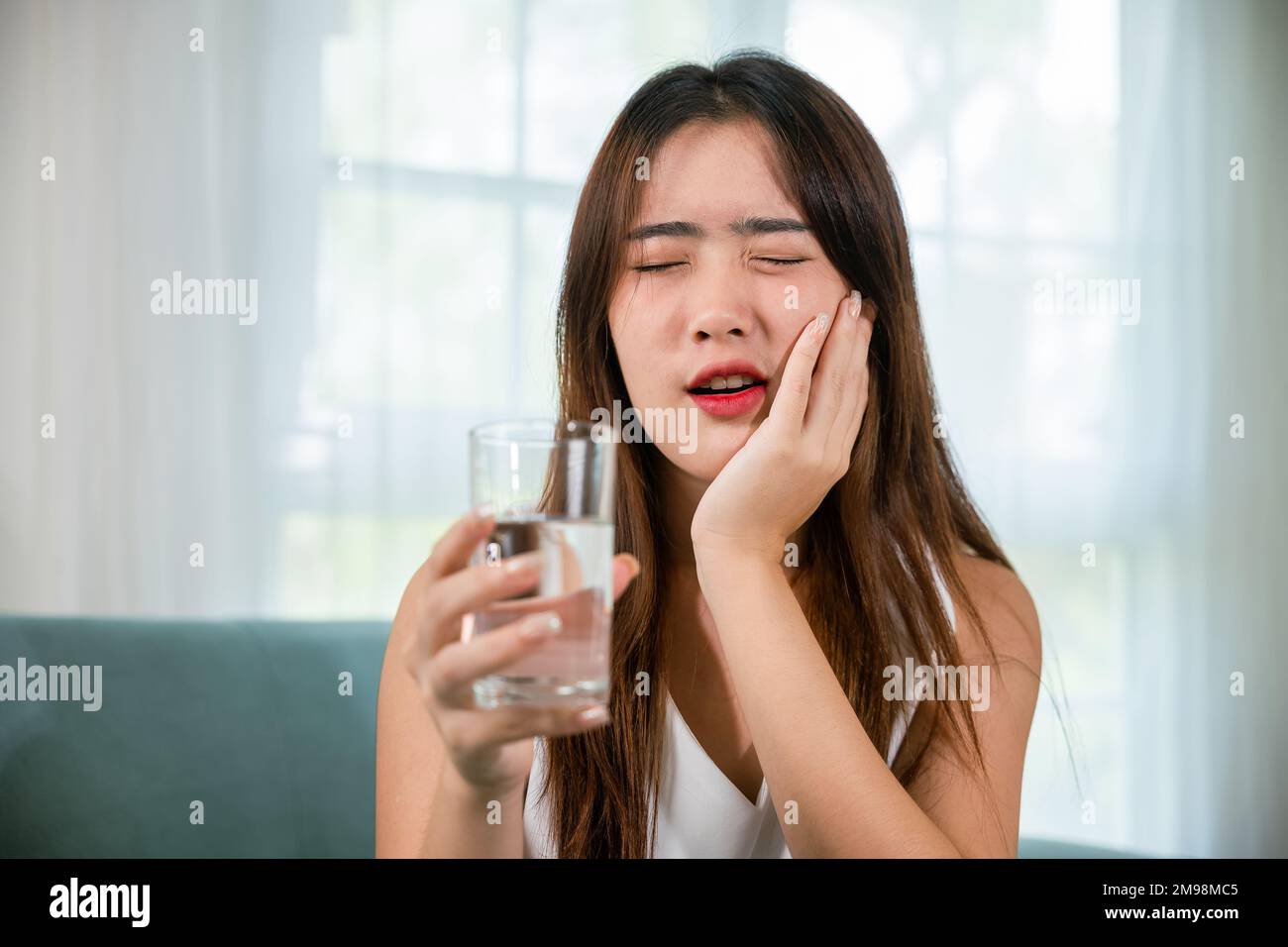 Giovane donna asiatica con denti sensibili che tiene un bicchiere d'acqua fredda Foto Stock