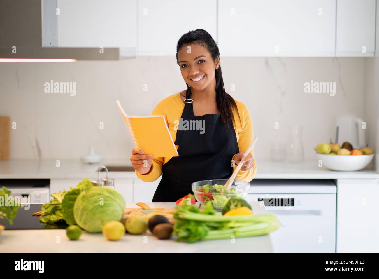 Allegra giovane afroamericana in grembiule preparare insalata e leggere ricettario Foto Stock