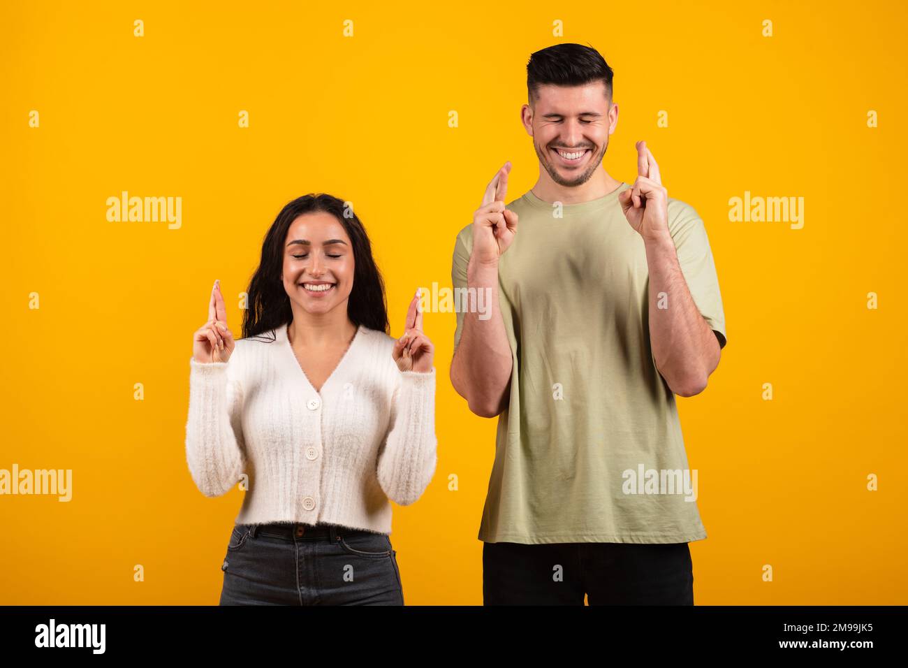 Felice ragazzo arabo millenario e femmina con gli occhi chiusi, le dita incrociate, fare un desiderio, isolato su sfondo arancione Foto Stock