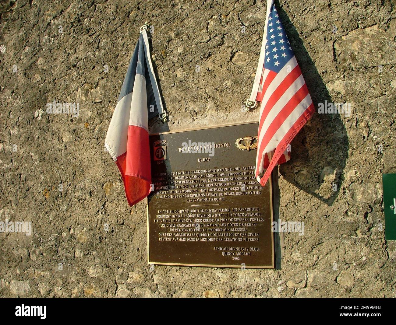 La lapide fu eretta dal 82nd Airborne C47 Club e commemora l'azione della 'Società D' del 2nd Battaglione 505 Paracute Infanteria Regiment, 82nd Divisione Airborne, nel cacciare un attacco tedesco contro Ste Mere Eglise. Riconosce anche l'aiuto dei francesi nel tendere i feriti. Dopo la Normandia le 505th insegne di spalla portarono il testo 'H-MENO' in basso perché il battaglione saltò prima di quanto avrebbe dovuto fare. Foto Stock