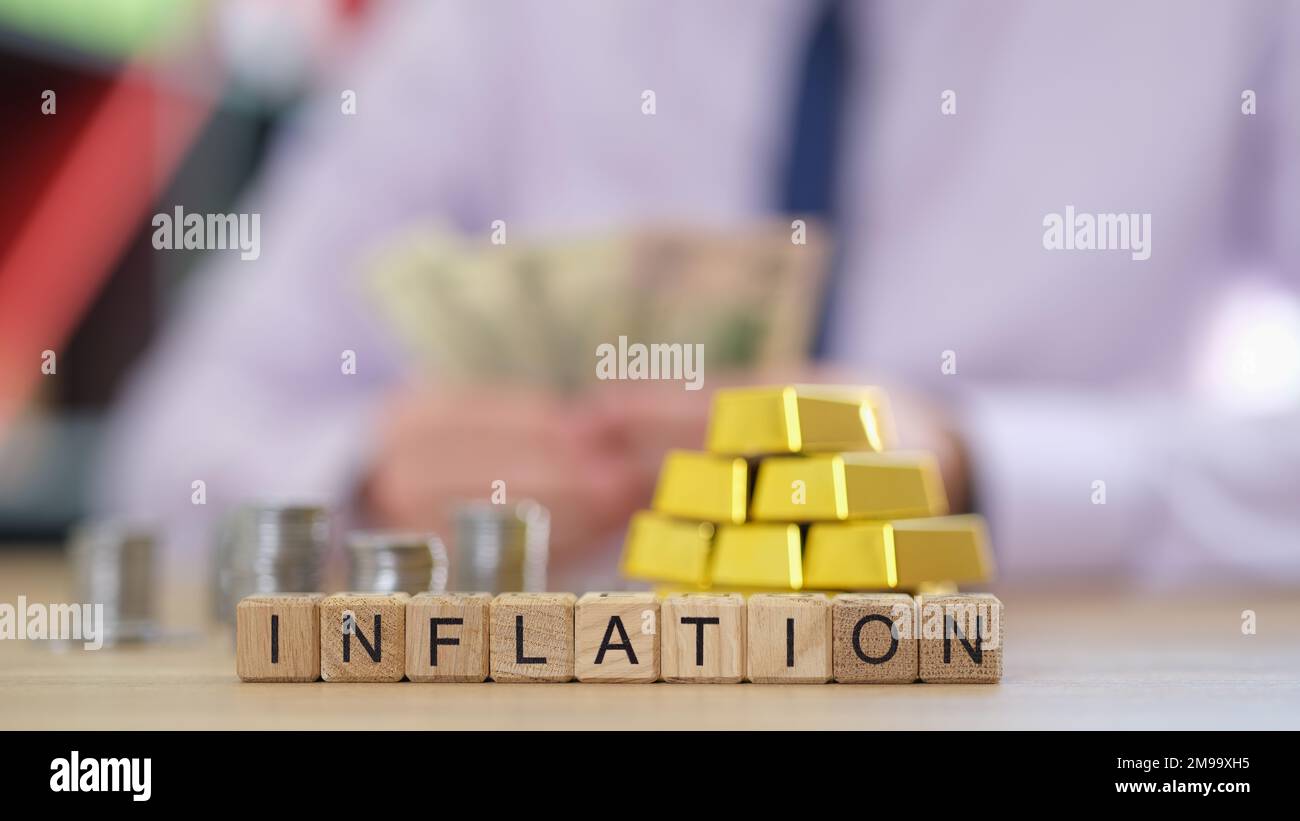 Lettere inflazione su cubi con barre d'oro e pile di monete, uomo d'affari conteggio di denaro in background. Foto Stock
