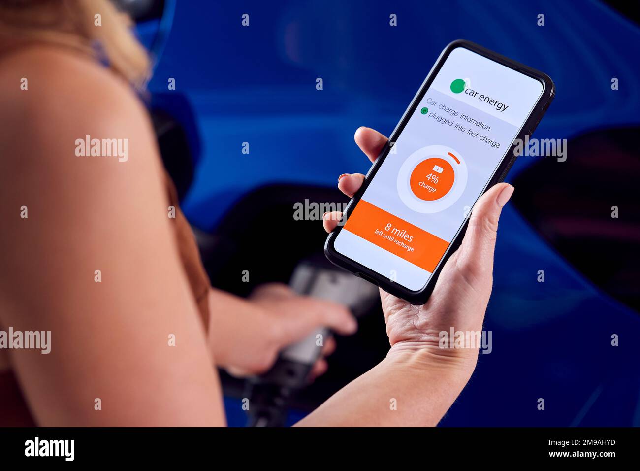Donna che carica l'auto elettrica con il cavo utilizzando l'app sul telefono per monitorare il livello della batteria Foto Stock