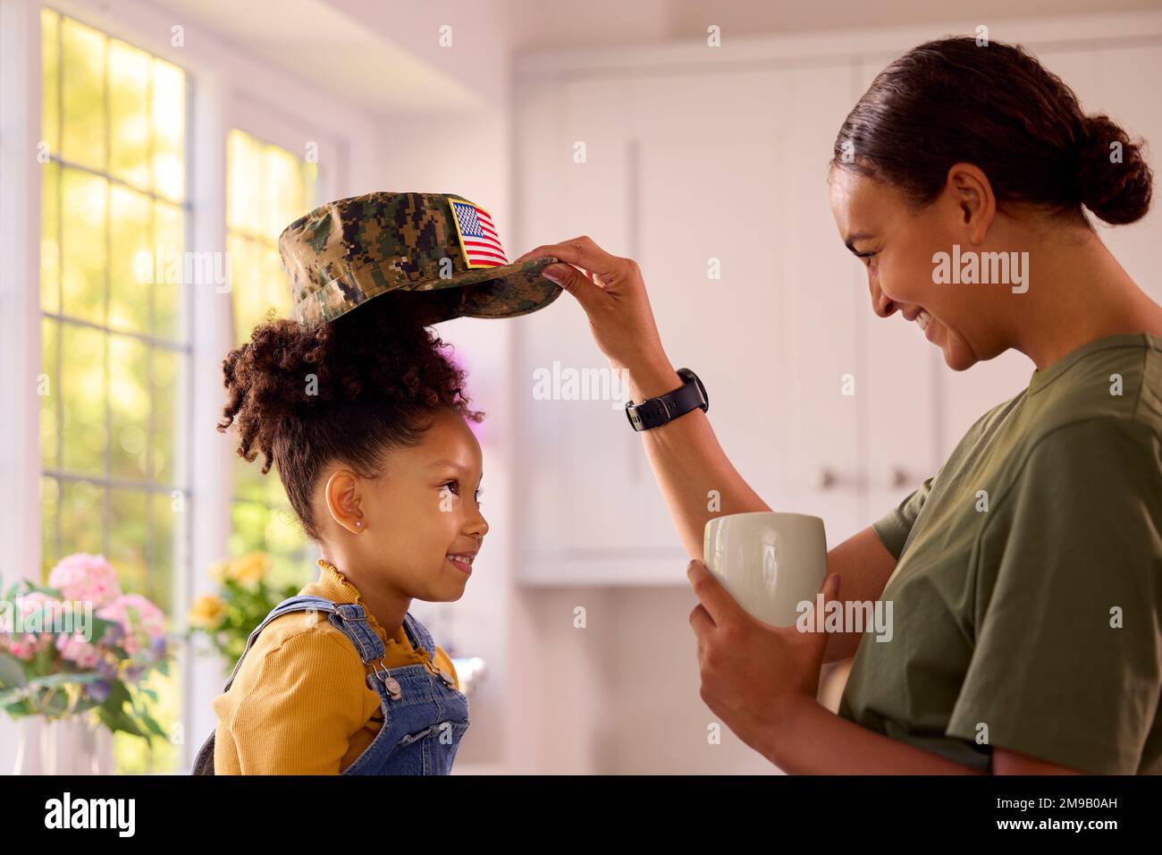 American Army Mother in uniforme Home on Leave mettere cappuccio su figlia seduta in cucina famiglia Foto Stock