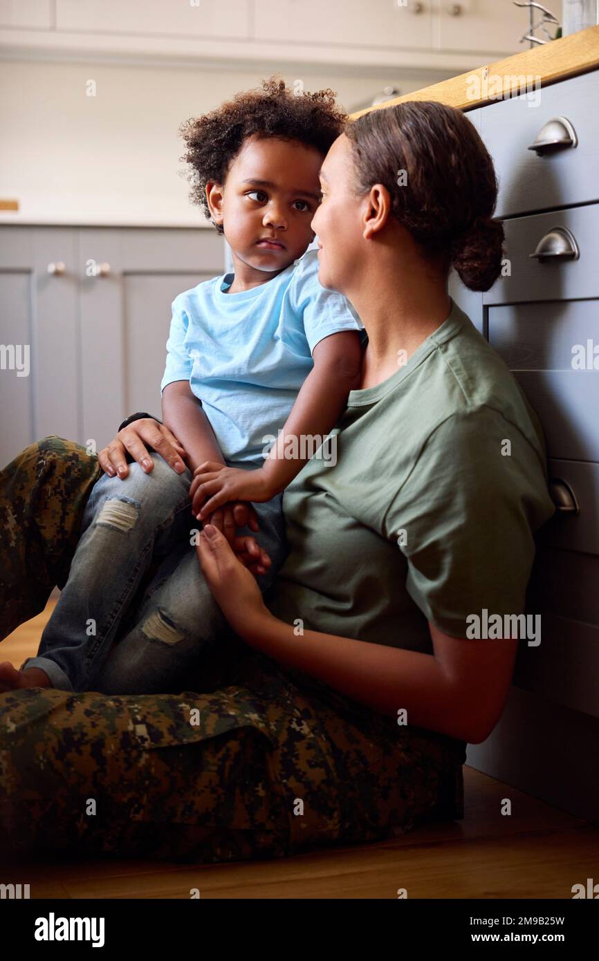 American Army Mother in uniforme Home on Leave Hugging Son Floor in Family Kitchen Foto Stock