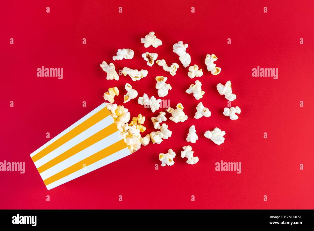 Gustoso popcorn al formaggio che cade da un secchio di cartone a strisce gialle, isolato su sfondo rosso. Dispersione di grani di popcorn. Film, cinema ed ente Foto Stock