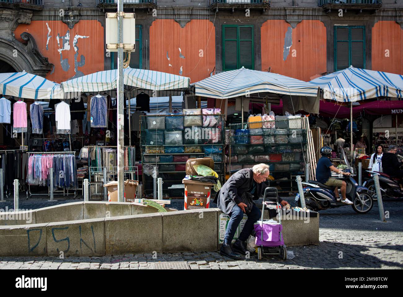 Un uomo si siede accanto a un mercato all'aperto nel quartiere di Sanita, Napoli. Ha un carrello per lo shopping viola Foto Stock