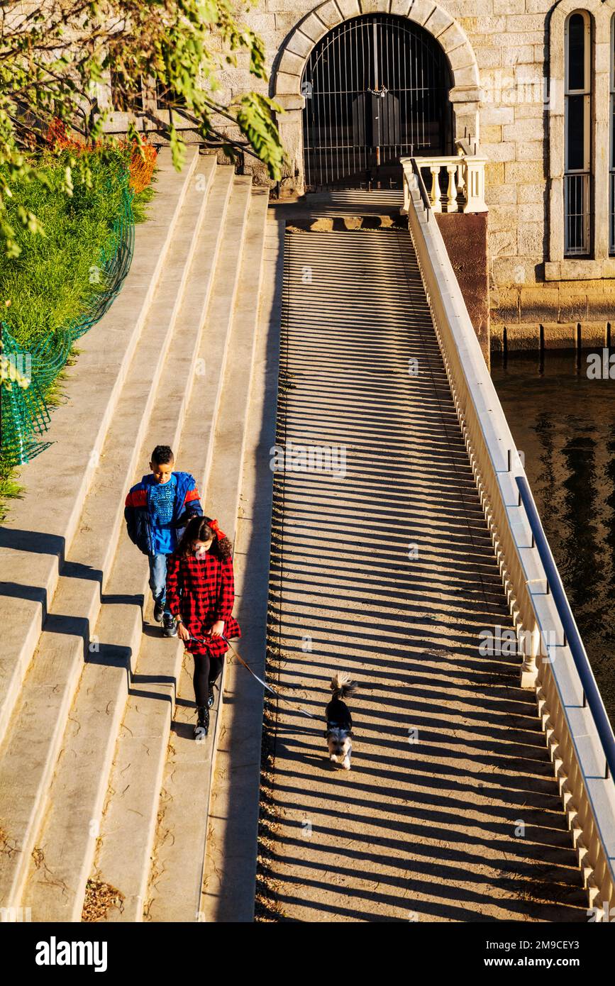 Giovane ragazzo e ragazza con cane da compagnia che saltano felicemente lungo il sentiero di pietra illuminato dal sole; Fairmount Water Works; Philadelphia; Pennsylvania; USA Foto Stock