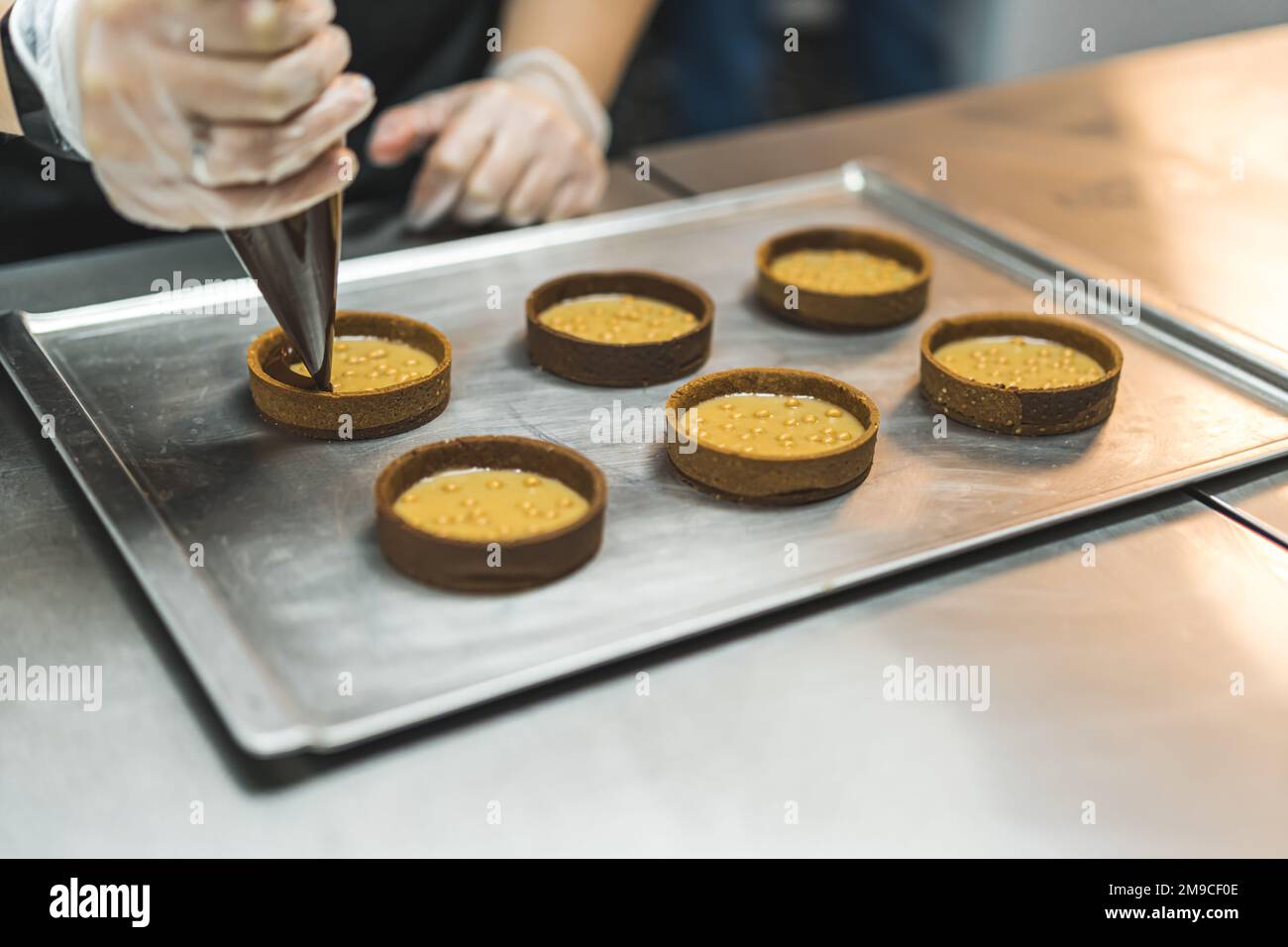 Panettiere irriconoscibile che colma ganache di cioccolato in croste di crostata mini riempite. Processo di cottura professionale. Assemblare dessert. Primo piano orizzontale in interni. Foto di alta qualità Foto Stock