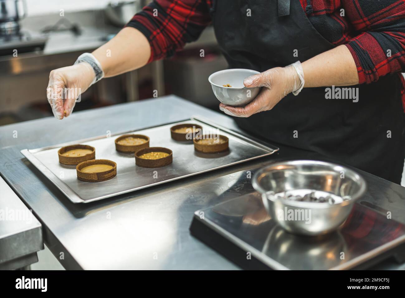 Panettiere professionale femminile che indossa grembiule nero cospargere dorate sulle croste crostate riempite. Processo di cottura. Assemblare dessert. Scatto orizzontale in interni. Foto di alta qualità Foto Stock