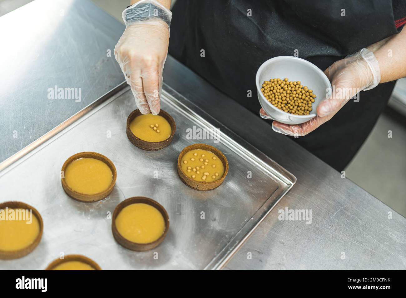 Vista dall'alto verso il basso del panettiere professionista che indossa un grembiule nero e spruzzi d'oro sulle piccole crostate riempite. Procedure di assemblaggio dei dessert. Scatto orizzontale in interni. Foto di alta qualità Foto Stock