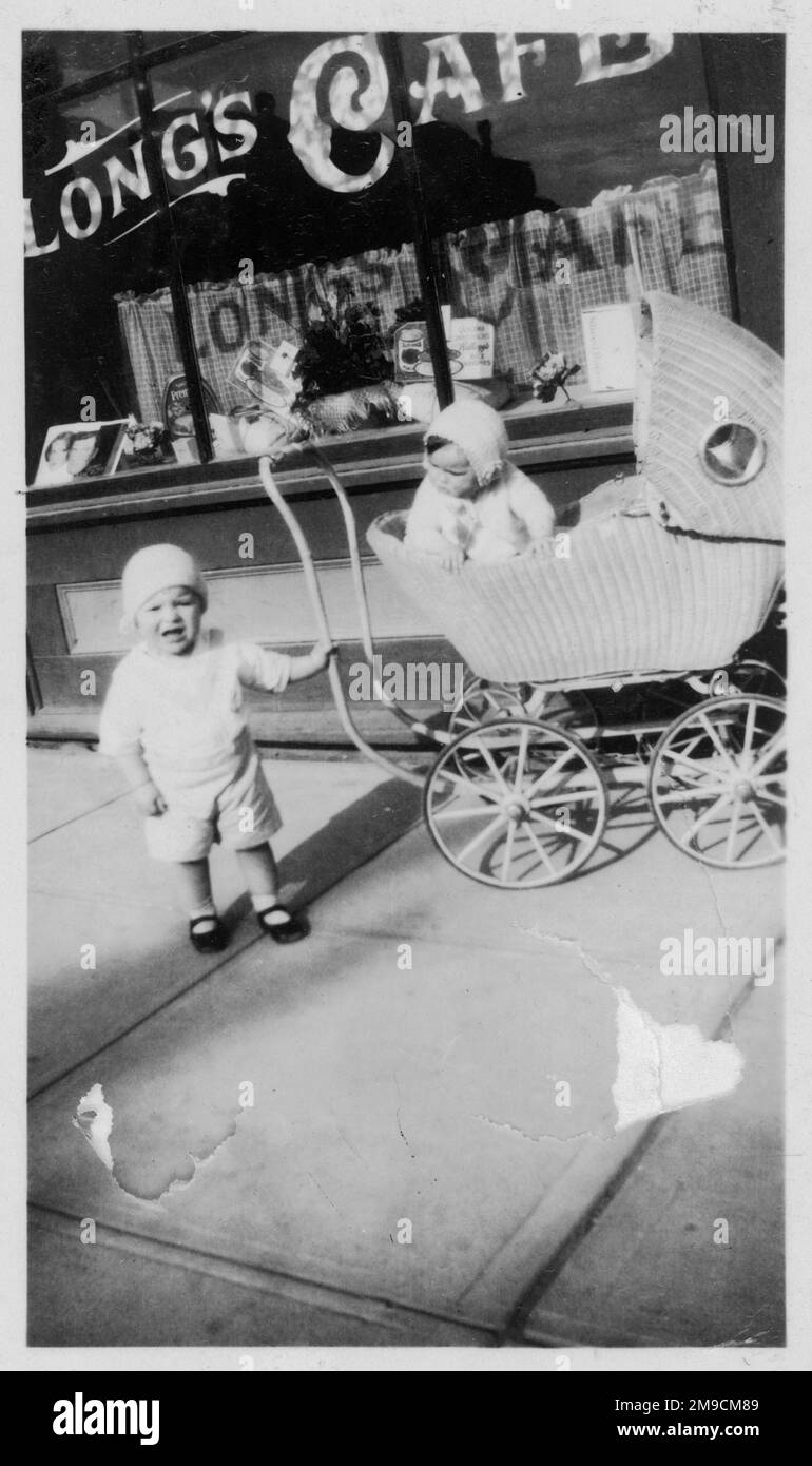 Un bambino che guarda infelice tiene il pram di sua sorella, mentre guarda giù a lui ansiosamente mentre aspettano la loro madre fuori del Long's Cafe. Foto Stock