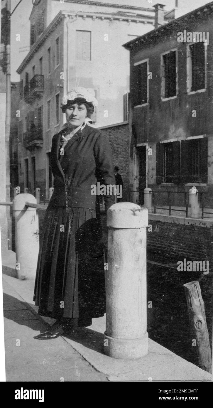 Donna in lungo vestito nero e cappello floreale si erge sul bordo di uno stretto canale a Venezia Foto Stock