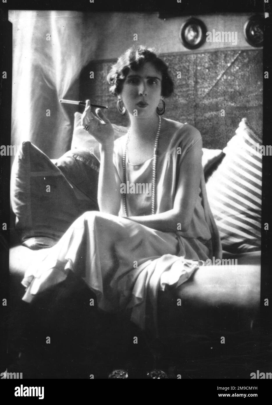Una donna dall'aspetto piuttosto imperioso che siede su un divano e fuma una sigaretta posta in un elegante supporto. Foto Stock