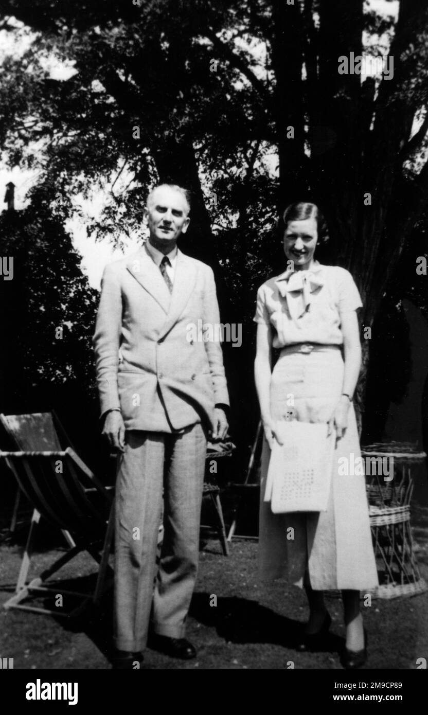 Elegante coppia inglese in piedi in un giardino vicino ad alcune sedie a sdraio, in parte all'ombra di un grande albero. Foto Stock