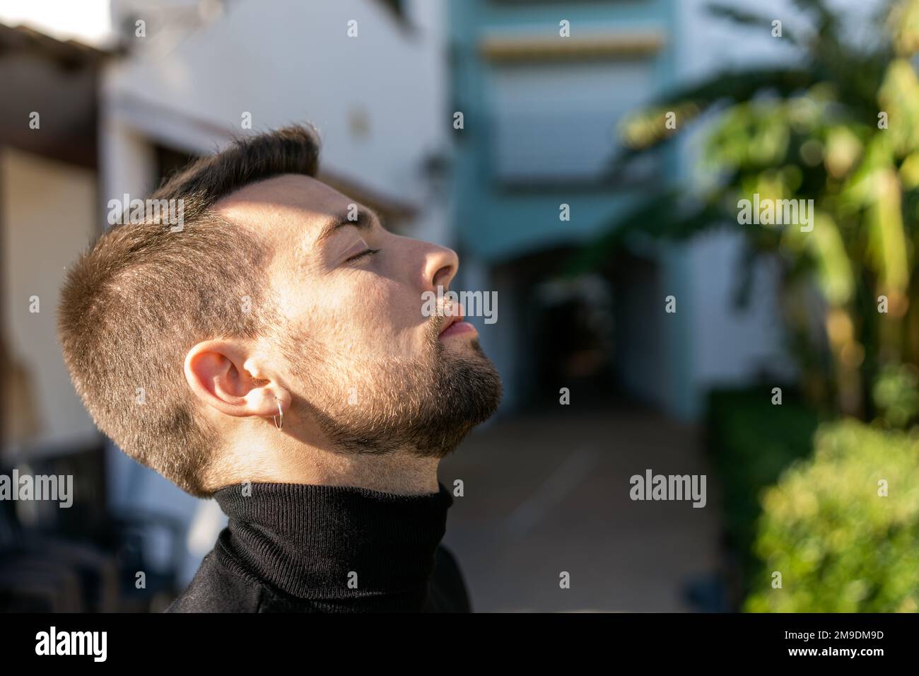 Vista laterale di un ragazzo calmo e bearded con orecchini e occhi chiusi che pende la testa indietro mentre si trova sullo sfondo sfocato della strada in condizioni di sole Foto Stock